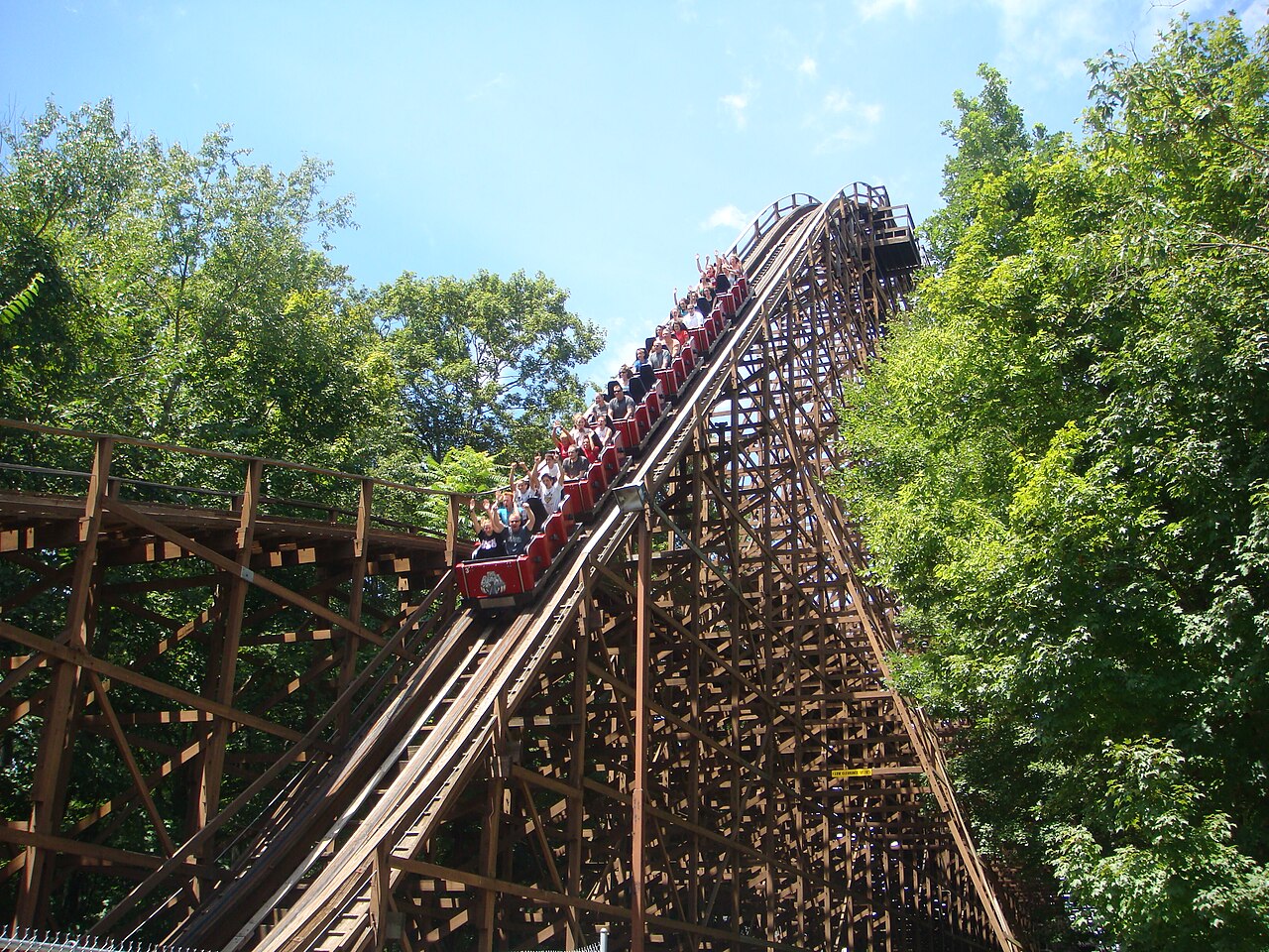 A red train full of riders climbing The Beast wooden track between tall trees
