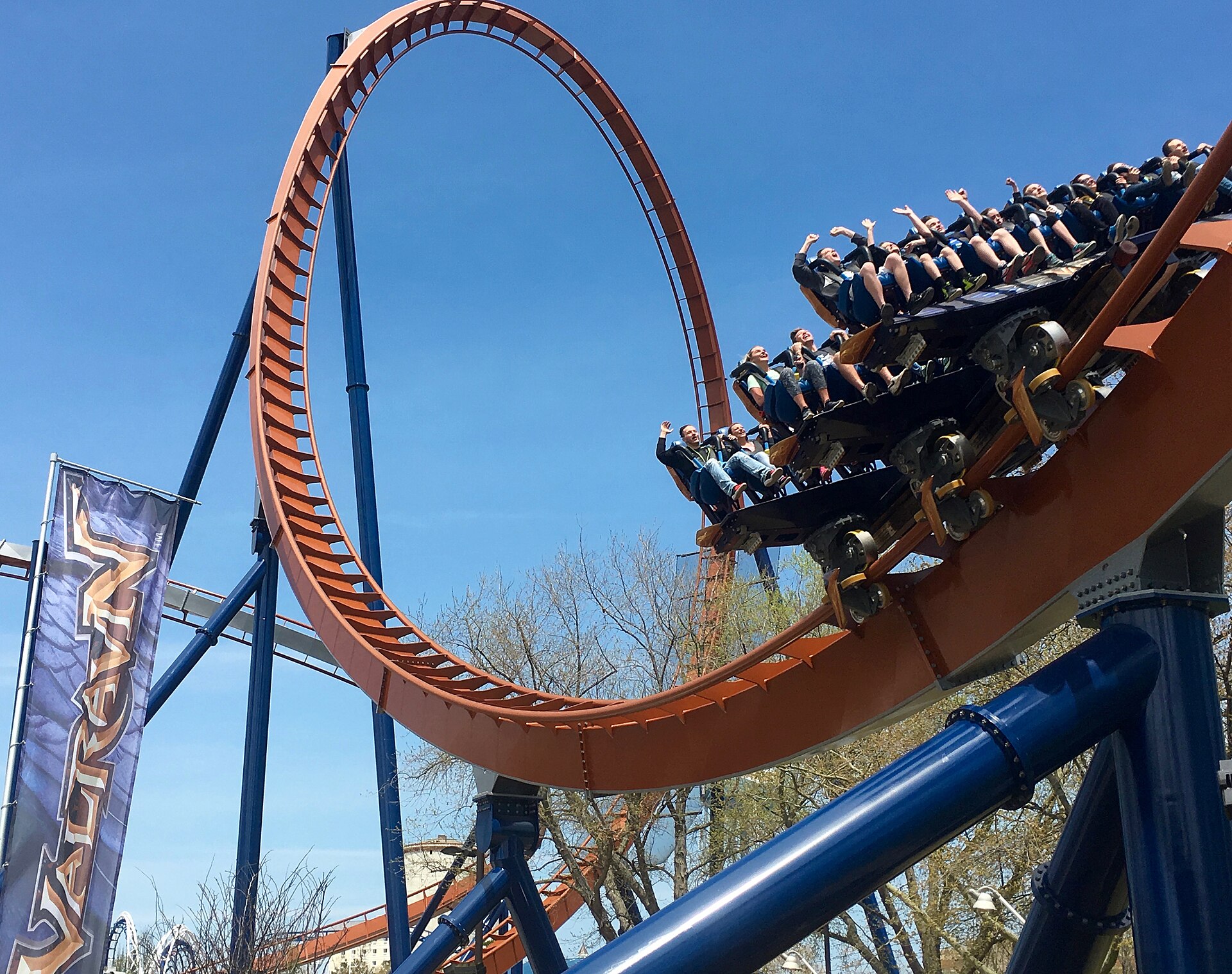 Riders with arms up on Valravn next to its orange loop under a blue sky