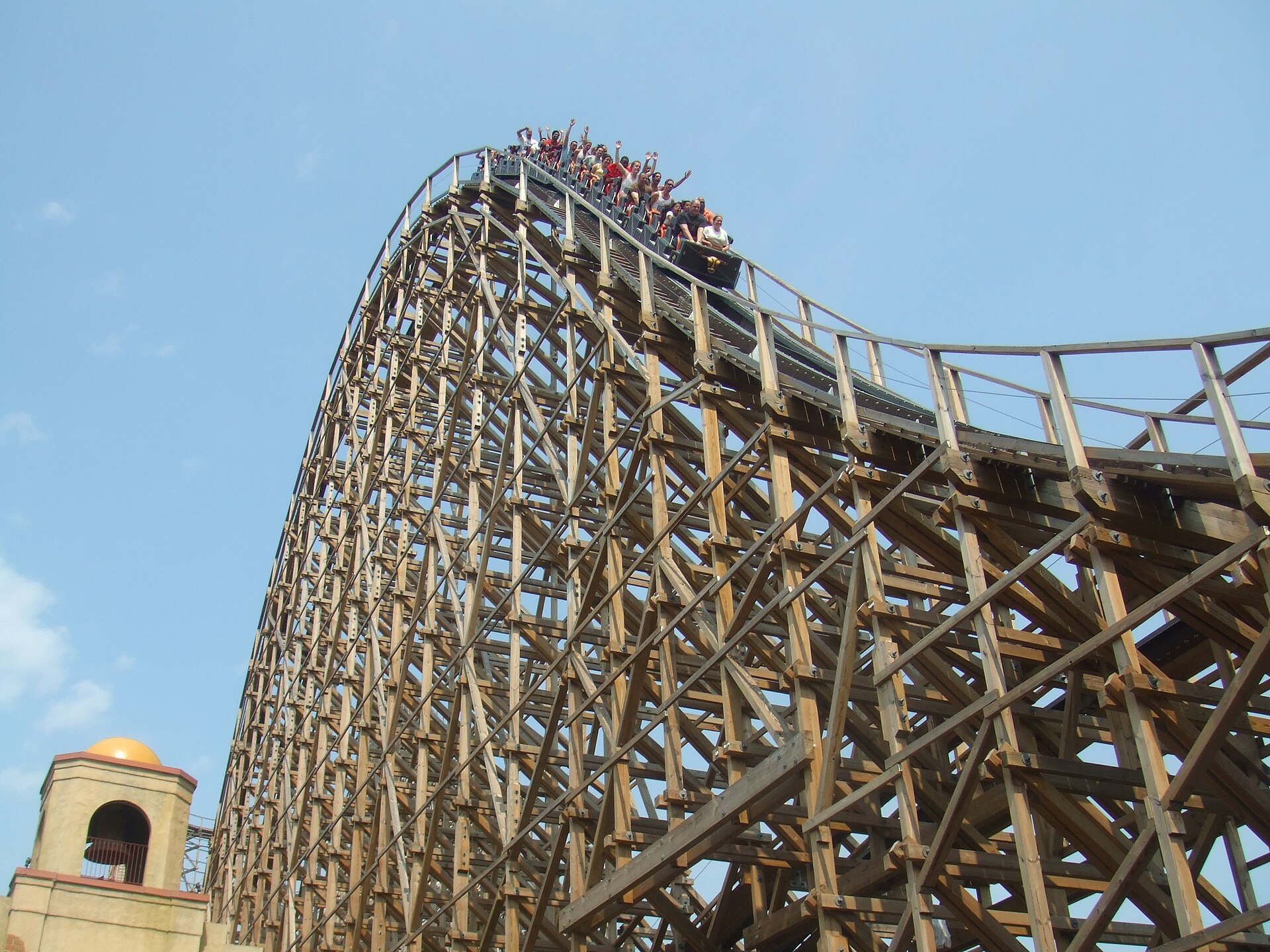 An El Toro train full of riders, arms up, cresting a hill on the tall wooden structure under a clear sky