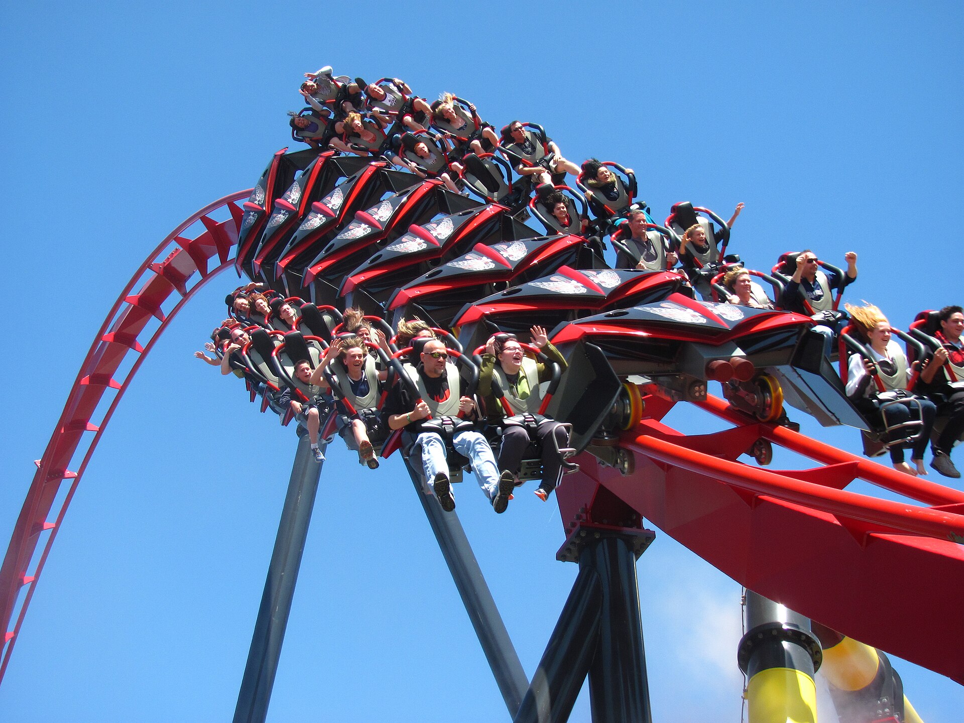 An X-Flight train full of riders, arms up, rolling out of an inversion on red track against a clear blue sky at Six Flags Great America
