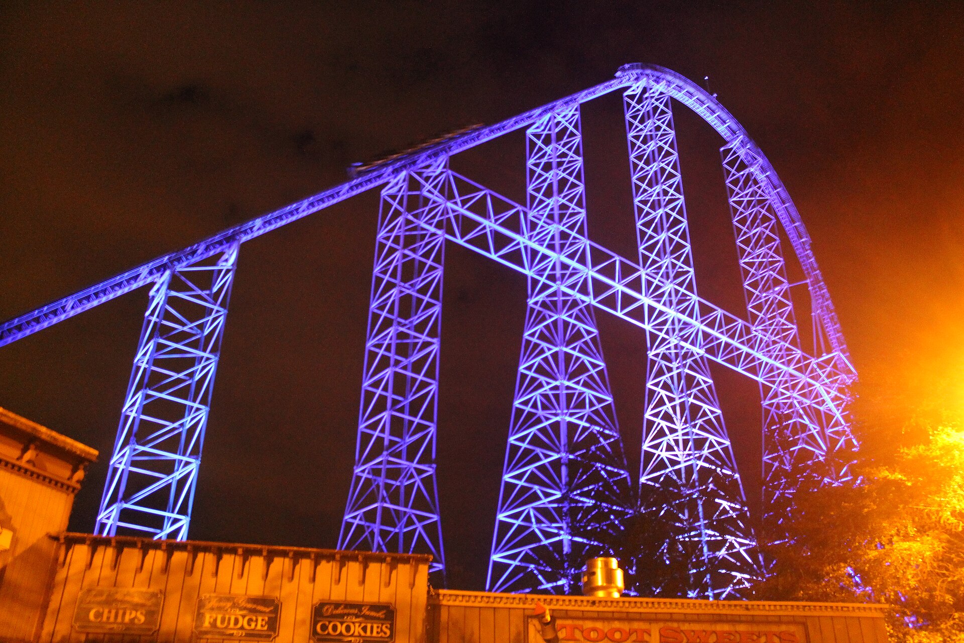A Millennium Force train cresting the top of the 310 ft blue lift hill above green summer trees, under a bright blue sky with white clouds